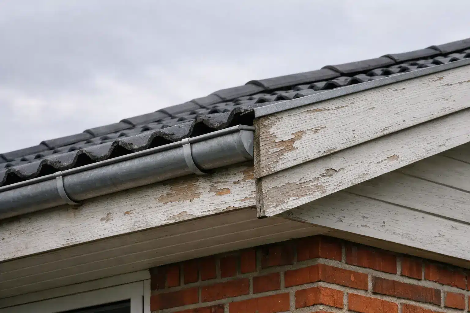 Foto af tagudhæng på dansk enfamiliehus med synlige sternbrædder og vindskeder. Træværket viser begyndende slid med afskallet maling og misfarvning, samt en let skæv tagrende i metal. Optaget i diffust dagslys under overskyet dansk vejr – velegnet til artikel om vedligeholdelse af tag og sternbrædder.