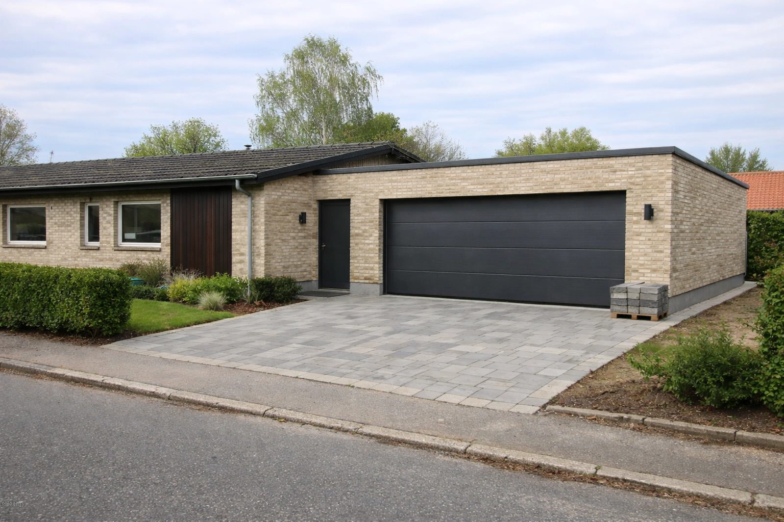 Moderniseret dansk 70’er parcelhus med integreret garagetilbygning, lyse mursten, mørk træbeklædning og antracit garageport.